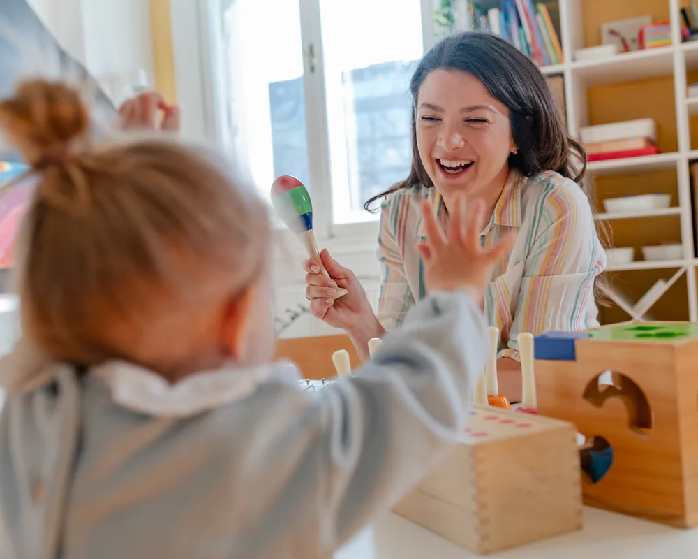 A toddler and a smiling educator playing with colourful maracas