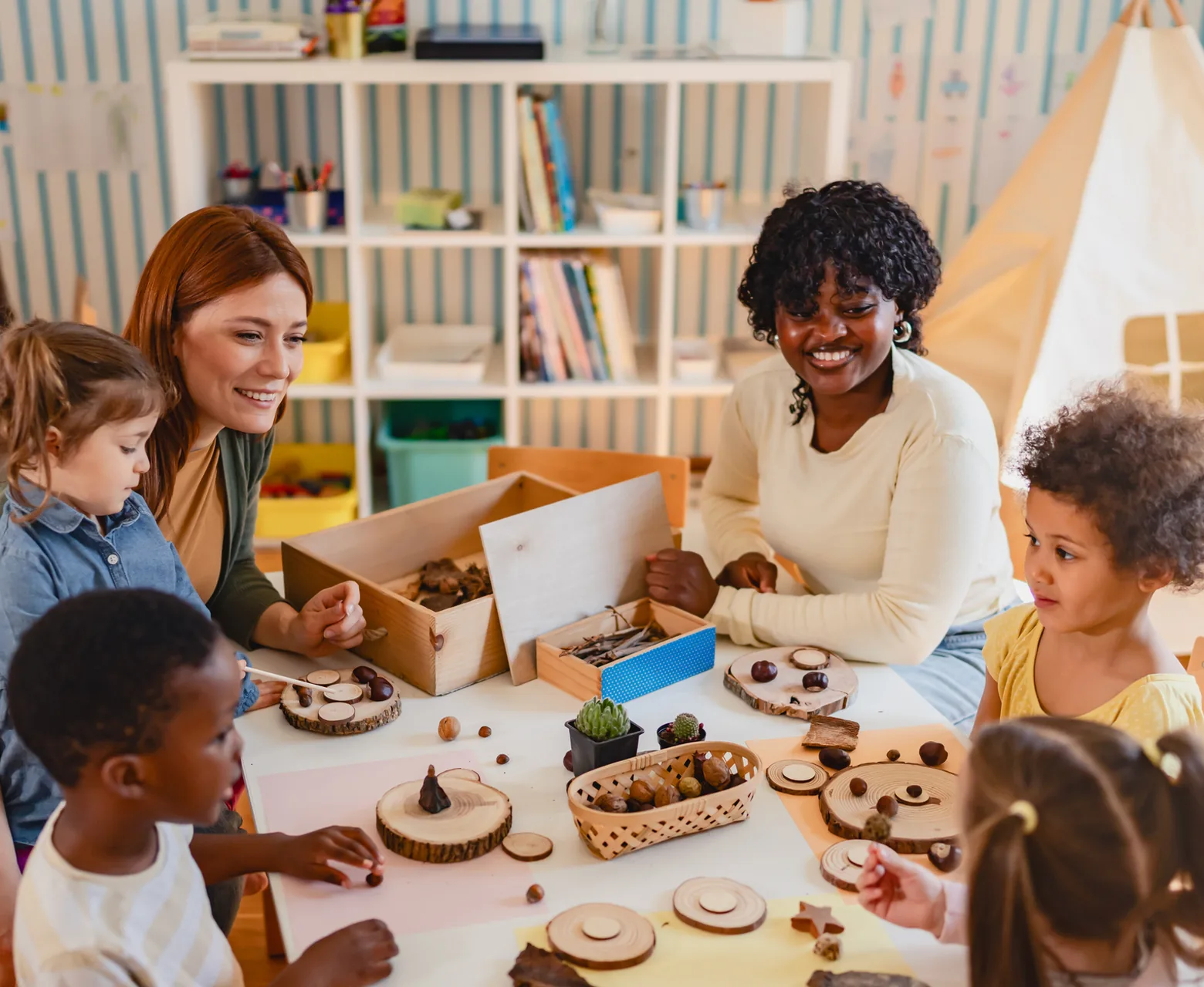 Two educators and a group of preschoolers exploring wooden trays of natural materials at a table